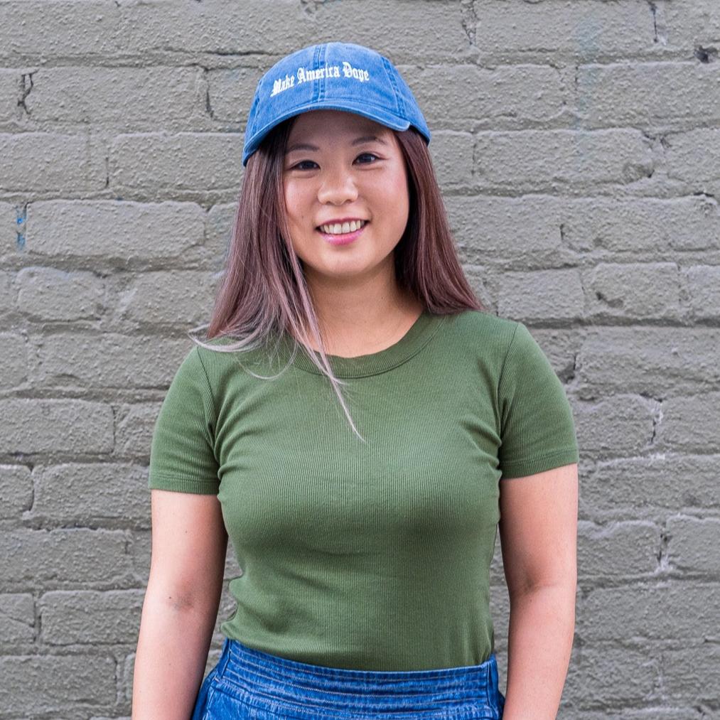Woman standing in front of olive wall wearing blue Make America Dope hat